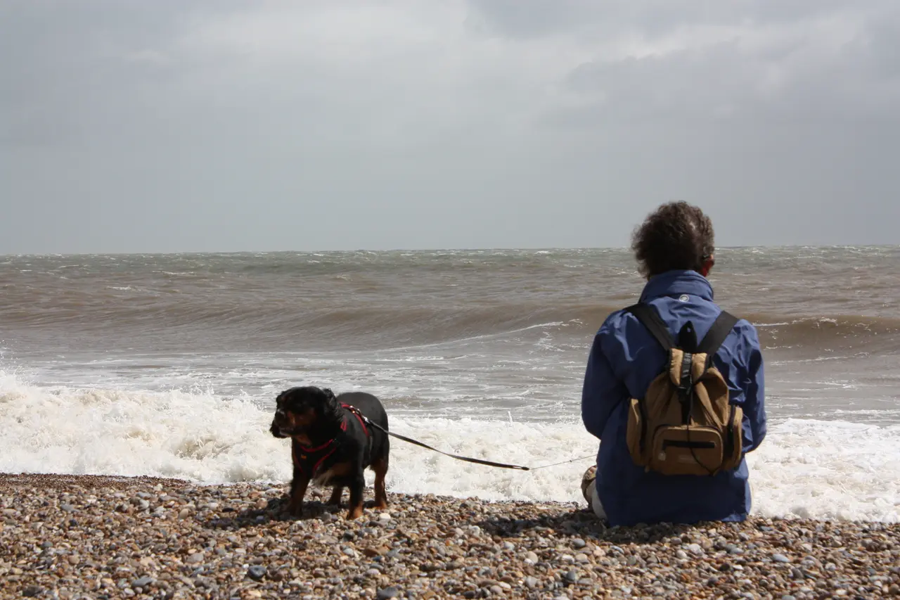 A person with a rucksack sitting on a shingle beach in Suffolk with a small black dog on a lead, waves breaking beyond